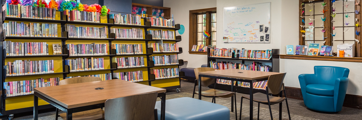 Photo of seating, tables, shelves, colorful decorations, and white board in the library teen and tween area