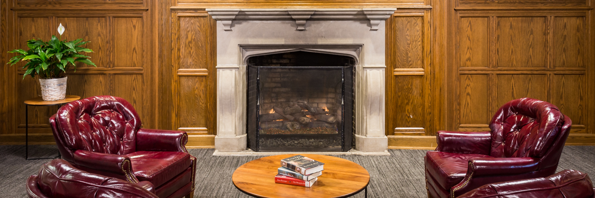 Reading area with stone fireplace, wood-paneled walls, wine-colored leather chairs, table with stack of books, and potted lily