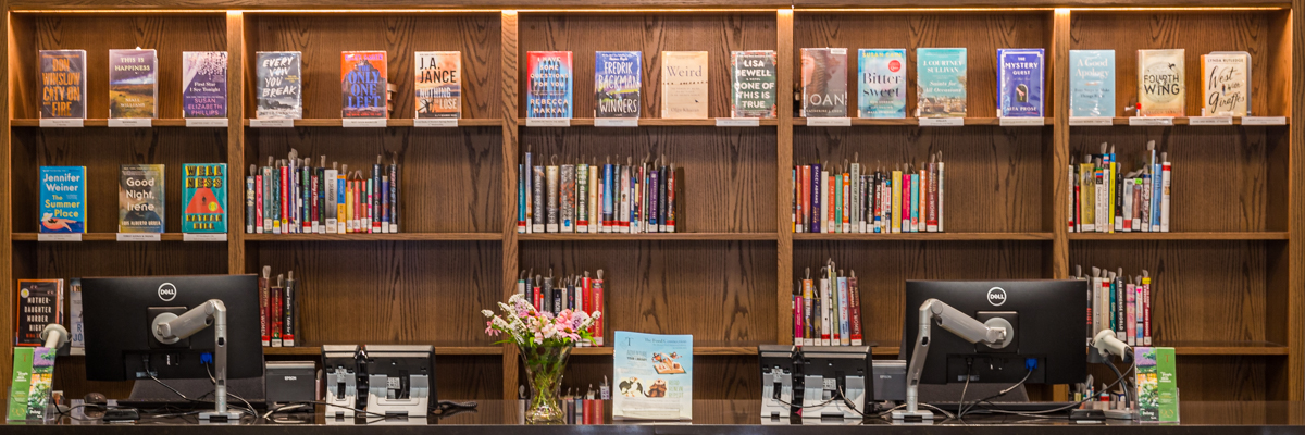 Library circulation desk with computer stations and wooden shelves displaying books behind