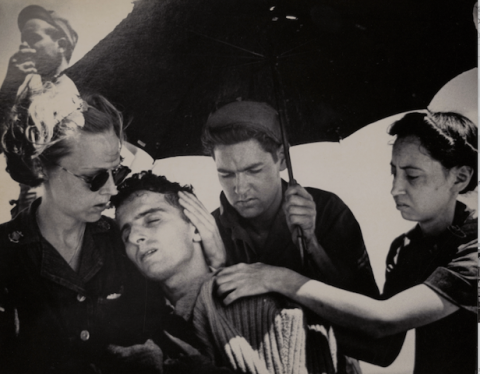 Two nurses caring for a soldier while another man holds an umbrella over them.